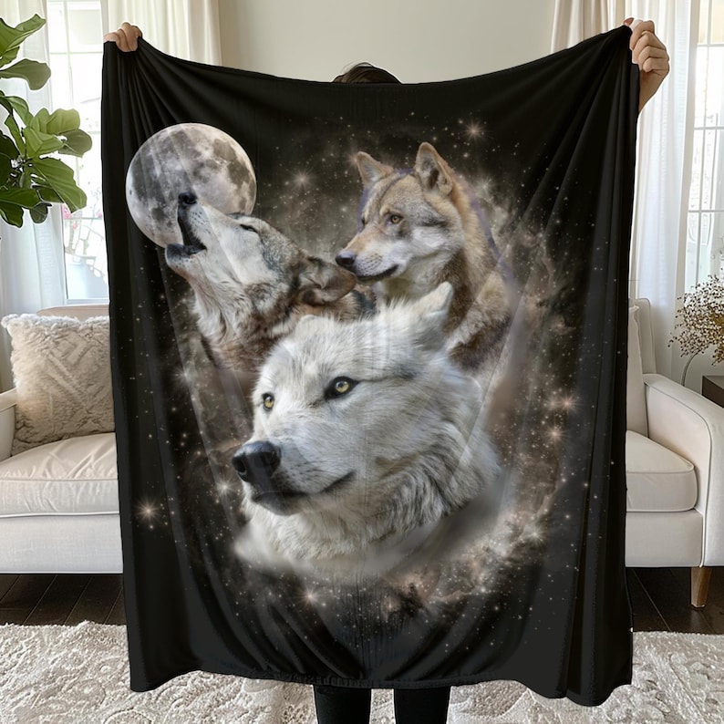 May include: Black blanket featuring a detailed illustration of three wolves under a full moon and starry night sky. The wolves are depicted in shades of white, brown, and gray, with glowing eyes. The blanket is held up by a person.