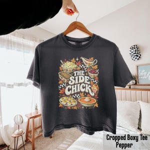 May include: A charcoal gray short-sleeve t-shirt featuring a colorful graphic print of various Thanksgiving side dishes, autumn leaves, and the text "THE SIDE CHICK" in a bold, retro-inspired font centered on the front of the garment.