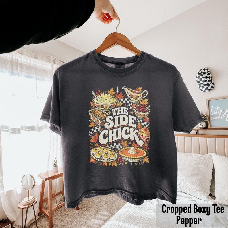 May include: A charcoal gray short-sleeve t-shirt featuring a colorful graphic print of various Thanksgiving side dishes, autumn leaves, and the text "THE SIDE CHICK" in a bold, retro-inspired font centered on the front of the garment.