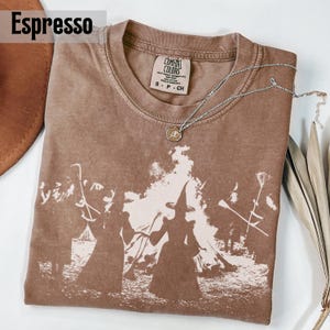 Pu&ograve; includere: Una t-shirt marrone a maniche corte con una stampa grafica bianca di figure con cappelli a punta vicino a un fal&ograve;. La maglia presenta un girocollo e un'etichetta Comfort Colors all'interno. Una collana d'argento &egrave; appoggiata sopra.