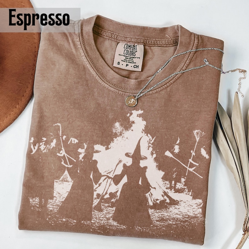 Pu&ograve; includere: Una t-shirt marrone a maniche corte con una stampa grafica bianca di figure con cappelli a punta vicino a un fal&ograve;. La maglia presenta un girocollo e un'etichetta Comfort Colors all'interno. Una collana d'argento &egrave; appoggiata sopra.