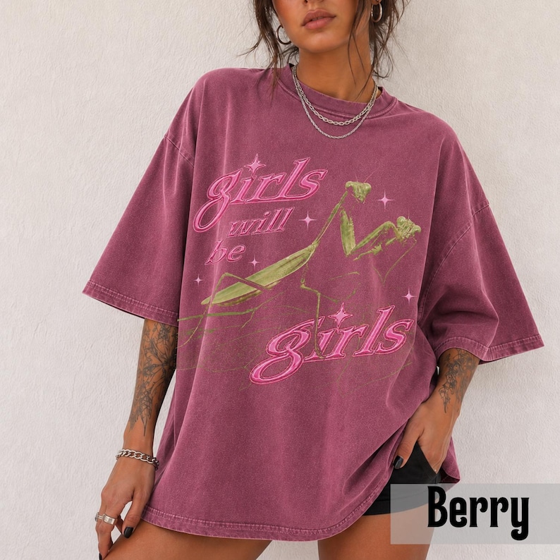 以下が含まれることがあります： ベリー色のオーバーサイズTシャツ。緑色のカマキリのグラフィックと、ピンクの筆記体で「Girls will be girls」という文字がプリントされています。クルーネック、半袖で、ヴィンテージ風のウォッシュ加工が施されています。