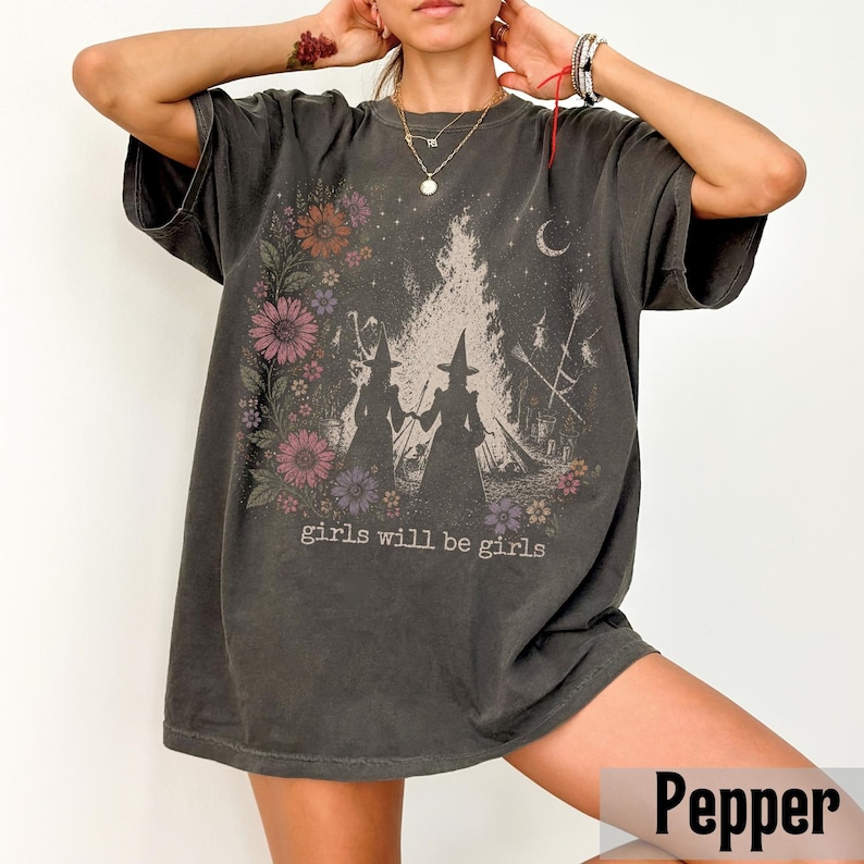 May include: Dark gray t-shirt with a graphic featuring two witch silhouettes, a bonfire, and floral accents. The text "girls will be girls" is printed below the graphic. The shirt has short sleeves and a relaxed fit.