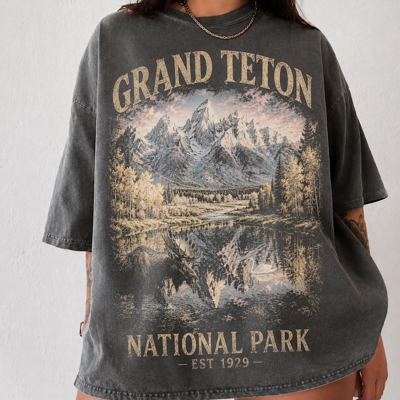 May include: Dark gray t-shirt featuring a scenic illustration of the Grand Teton National Park. The design includes mountains, a lake with a moose, and the text "GRAND TETON NATIONAL PARK - EST 1929-". The shirt has short sleeves and a relaxed fit.