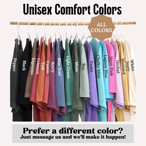 May include: A display of various colored t-shirts hanging on a wooden rod. The shirts are in shades like black, crimson, and lagoon blue. Text on the image reads "Unisex Comfort Colors" and "All Colors."
