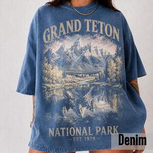May include: Denim blue t-shirt featuring a scenic illustration of Grand Teton National Park. The design includes a mountain range, a lake, and trees, with the text "GRAND TETON NATIONAL PARK - EST 1929".