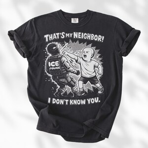 K&ouml;nnte beinhalten: Schwarzes T-Shirt mit einer Grafik eines Babys, das einen ICE-Polizisten schl&auml;gt. Der Text auf dem Shirt lautet: "THAT'S MY NEIGHBOR! I DON'T KNOW YOU."