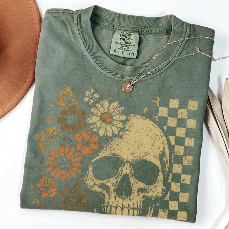 May include: Olive green Comfort Colors t-shirt featuring a skull design with floral accents and a checkered pattern. A silver necklace with a pendant rests on the shirt. The shirt is folded and ready to wear.