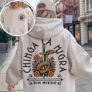 May include: Cream-colored hoodie with a graphic on the back featuring a snake coiled around a sword, surrounded by flowers. The text "CHINGA LA MIGRA" and "SIN MIEDO" are also displayed. A smaller version of the graphic is on the front.