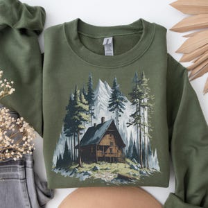 May include: Olive green sweatshirt featuring a mountain cabin scene. The graphic depicts a wooden cabin nestled among tall pine trees and snow-capped mountains. The design is printed on a soft, crew neck sweatshirt.