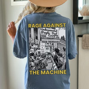 Puede incluir: Camiseta azul claro de manga corta con un gr&aacute;fico en la espalda. El gr&aacute;fico presenta el texto "RAGE AGAINST THE MACHINE" en amarillo, junto con una imagen en blanco y negro de una protesta. La imagen incluye el texto "Undocumented People Feed Everyone BILLIONAIRES".