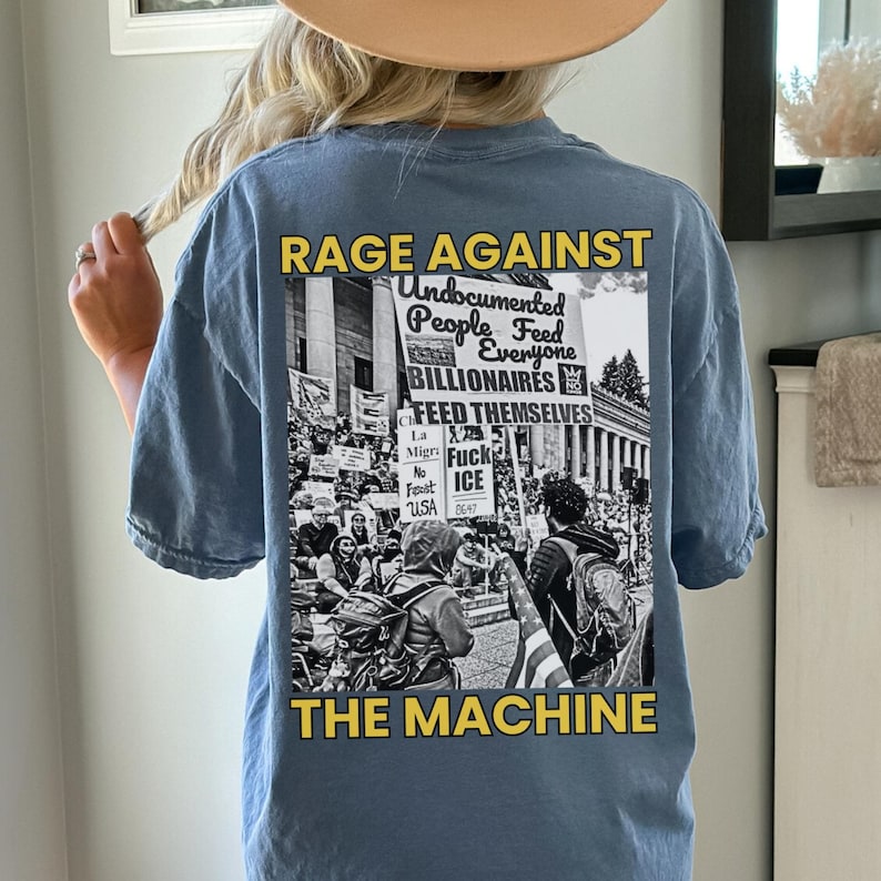 Puede incluir: Camiseta azul claro de manga corta con un gr&aacute;fico en la espalda. El gr&aacute;fico presenta el texto "RAGE AGAINST THE MACHINE" en amarillo, junto con una imagen en blanco y negro de una protesta. La imagen incluye el texto "Undocumented People Feed Everyone BILLIONAIRES".