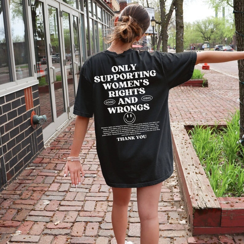 May include: Black t-shirt with white text that reads "Only Supporting Women's Rights and Wrongs." The text is surrounded by a smiley face and the words "Good Vibes".