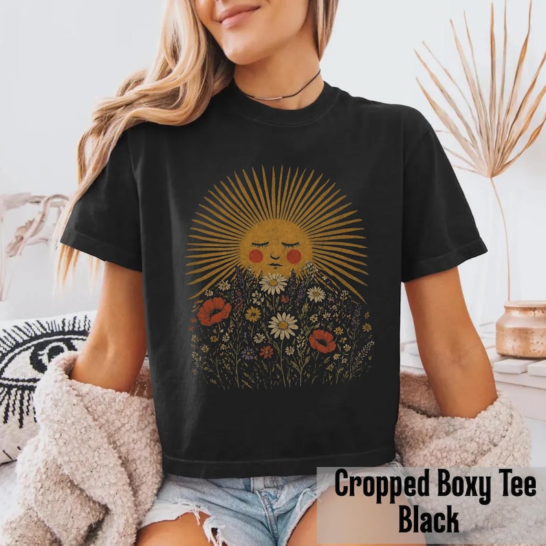 Puede incluir: Camiseta negra corta y holgada con un gr&aacute;fico de sol con cara, rodeado de flores. El sol tiene rayos dorados y una cara roja. Las flores incluyen amapolas rojas y margaritas blancas. El texto "Cropped Boxy Tee Black" est&aacute; en la parte inferior.