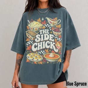 May include: A dark gray oversized t-shirt featuring a graphic print of various Thanksgiving side dishes, including deviled eggs, a pie, and a gravy boat. The text reads THE SIDE CHICK in large white letters surrounded by autumn leaves.