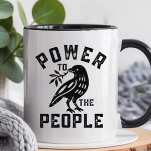 May include: White ceramic mug with a black rim and handle. The mug features the text "POWER TO THE PEOPLE" in bold, black letters, along with a black illustration of a bird holding a sprig. The mug is set against a blurred background.