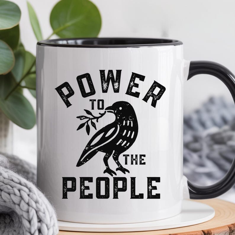 May include: White ceramic mug with a black rim and handle. The mug features the text "POWER TO THE PEOPLE" in bold, black letters, along with a black illustration of a bird holding a sprig. The mug is set against a blurred background.