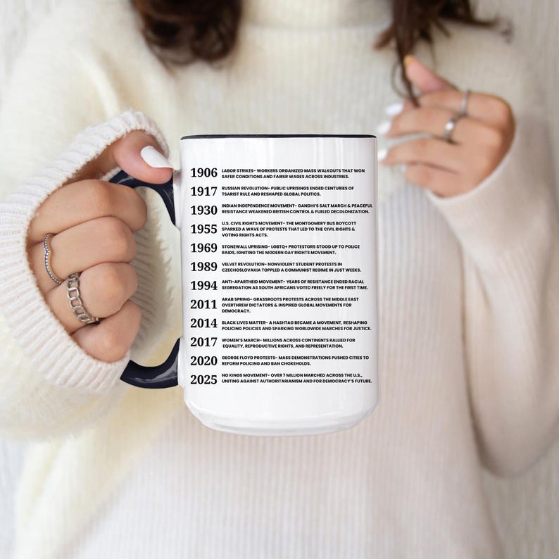 May include: A white ceramic mug with a black interior and handle, held by a person. The mug features a timeline of historical events, including labor strikes, civil rights, and women's marches. The text is in black.