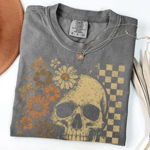 May include: A gray Comfort Colors t-shirt featuring a skull graphic with floral and checkered elements in shades of gold and orange. A silver necklace with a pendant rests on the shirt.