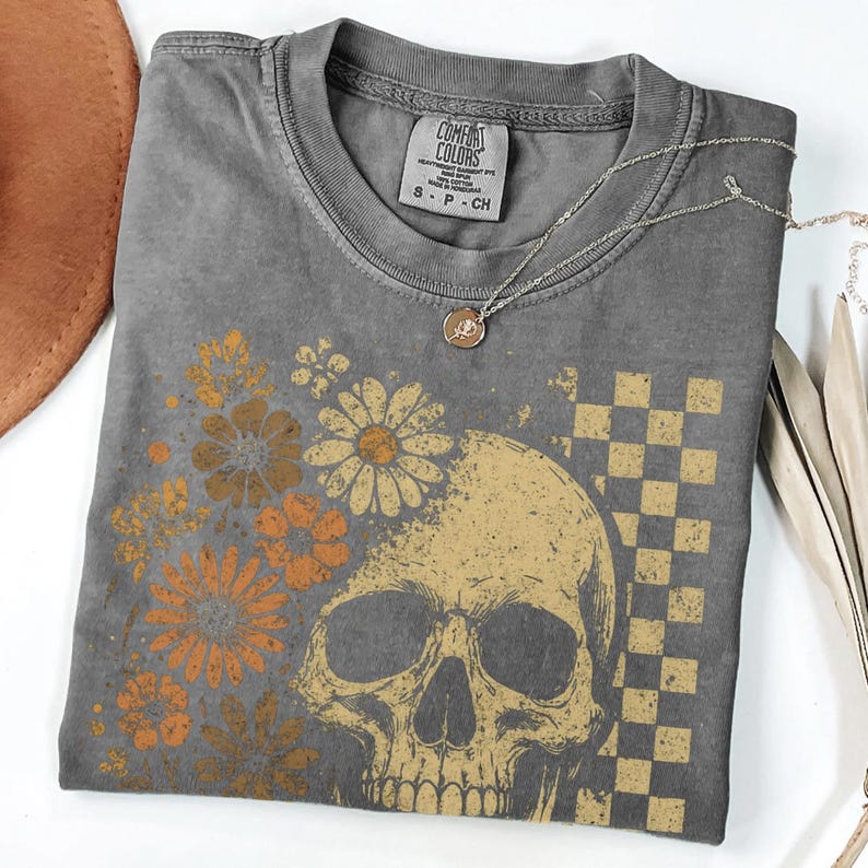 May include: A gray Comfort Colors t-shirt featuring a skull graphic with floral and checkered elements in shades of gold and orange. A silver necklace with a pendant rests on the shirt.