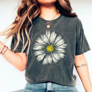 May include: A gray t-shirt with a white daisy graphic and the text "Find the good and hold on" printed around the daisy. The shirt has a relaxed fit and short sleeves.