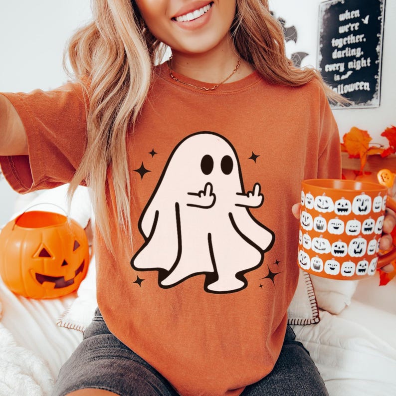 May include: Orange t-shirt featuring a cartoon ghost giving the middle finger, with black star accents. The image also includes an orange pumpkin-shaped container and a mug with jack-o'-lantern faces, perfect for Halloween.