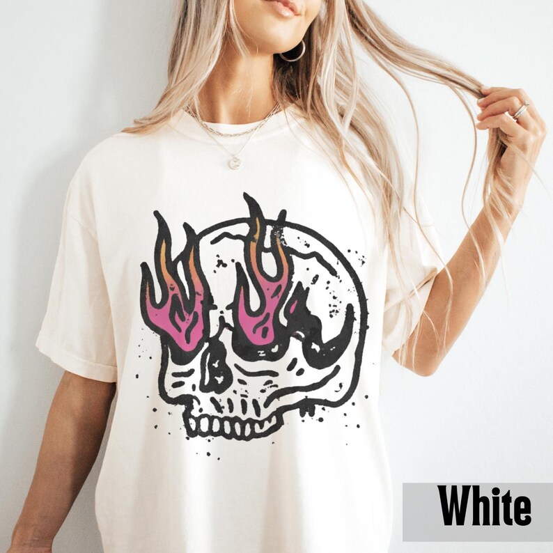 May include: White t-shirt featuring a black and white skull graphic with flames in pink and orange. The shirt has a relaxed fit and short sleeves. The word "White" is printed in a white box at the bottom right.