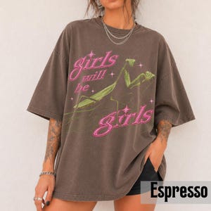 以下が含まれることがあります： エスプレッソカラーのオーバーサイズTシャツ。緑色のカマキリのグラフィックが描かれています。ピンク色の文字で「girls will be girls」とプリントされており、星のアクセントが添えられています。クルーネックで、袖は短くゆったりとした作りです。