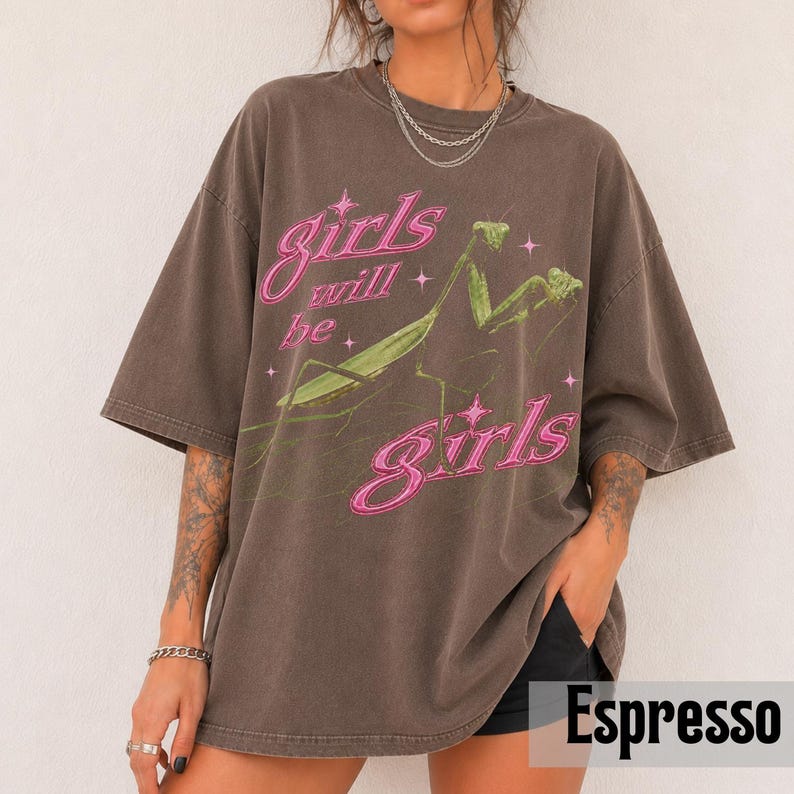 以下が含まれることがあります： エスプレッソカラーのオーバーサイズTシャツ。緑色のカマキリのグラフィックが描かれています。ピンク色の文字で「girls will be girls」とプリントされており、星のアクセントが添えられています。クルーネックで、袖は短くゆったりとした作りです。