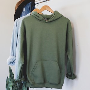 May include: An olive green hoodie with a front pocket hangs on a wooden hanger. A light blue long-sleeve shirt and a brown hat are also on the rack. The hoodie is made of a soft material and has a hood.
