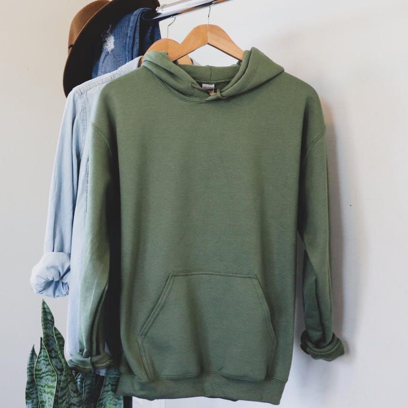 May include: An olive green hoodie with a front pocket hangs on a wooden hanger. A light blue long-sleeve shirt and a brown hat are also on the rack. The hoodie is made of a soft material and has a hood.