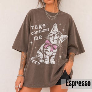 May include: A dark brown oversized t-shirt featuring a graphic of a tabby kitten wearing a pink bow. The text "rage consumes me" is printed in white above the kitten, surrounded by small white star accents. The shirt has a crew neckline.
