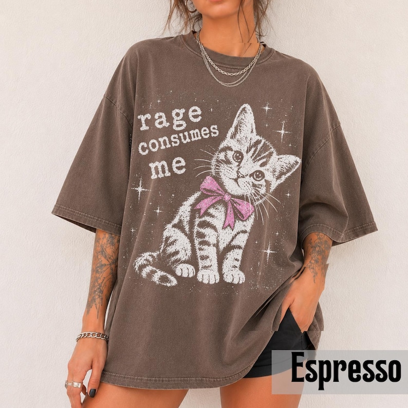 May include: A dark brown oversized t-shirt featuring a graphic of a tabby kitten wearing a pink bow. The text "rage consumes me" is printed in white above the kitten, surrounded by small white star accents. The shirt has a crew neckline.