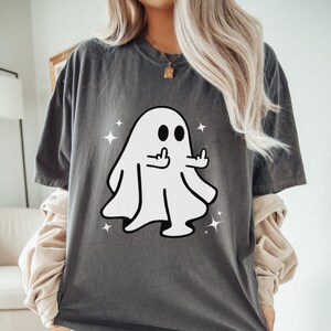 May include: Dark gray t-shirt featuring a white cartoon ghost giving the middle finger. The ghost has black eyes and is surrounded by white star accents. The shirt is a casual style.