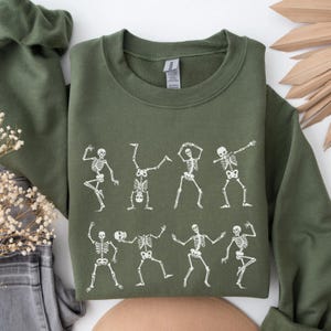 K&ouml;nnte beinhalten: Ein olivgr&uuml;nes Sweatshirt mit einer wei&szlig;en Grafik von tanzenden Skeletten in verschiedenen Posen. Das Design zeigt Skelette, die Handst&auml;nde machen und andere Bewegungen. Das Sweatshirt ist aus weichem Material.