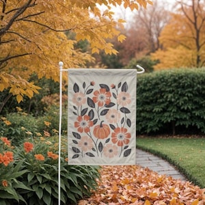 Puede incluir: Una bandera de jard&iacute;n beige con un dise&ntilde;o floral de flores naranjas y melocot&oacute;n, hojas grises y una calabaza. La bandera se muestra en un soporte de metal blanco en un jard&iacute;n con follaje oto&ntilde;al.