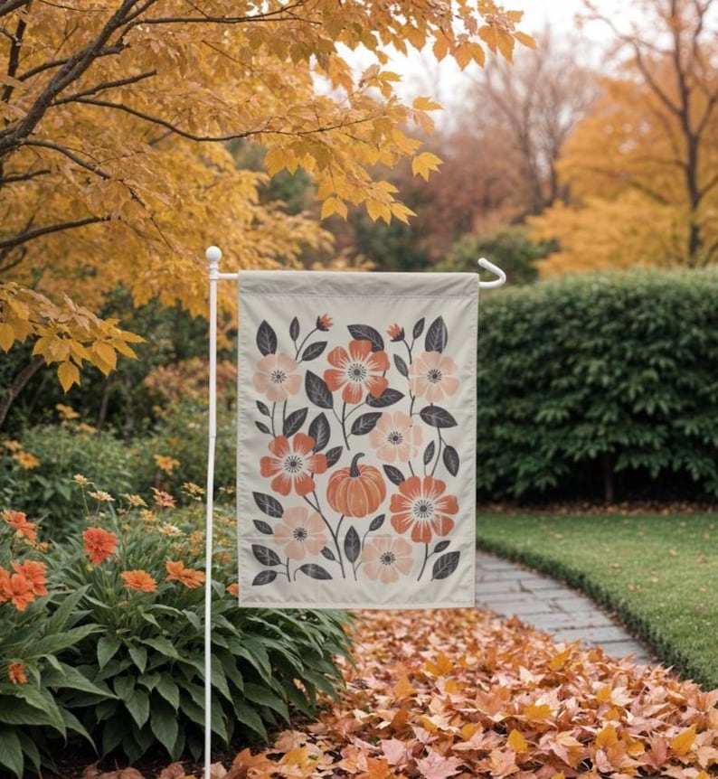 Puede incluir: Una bandera de jard&iacute;n beige con un dise&ntilde;o floral de flores naranjas y melocot&oacute;n, hojas grises y una calabaza. La bandera se muestra en un soporte de metal blanco en un jard&iacute;n con follaje oto&ntilde;al.