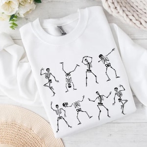 K&ouml;nnte beinhalten: Wei&szlig;es Sweatshirt mit einem schwarzen Aufdruck tanzender Skelette. Die Skelette sind in verschiedenen Posen dargestellt, mit ausgestreckten Armen und Beinen. Das Design befindet sich mittig auf der Vorderseite des Sweatshirts.