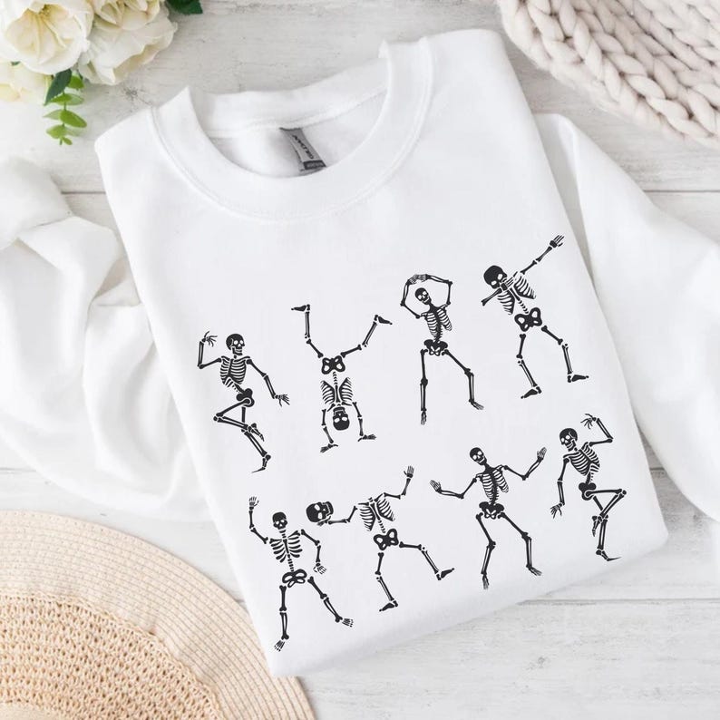 K&ouml;nnte beinhalten: Wei&szlig;es Sweatshirt mit einem schwarzen Aufdruck tanzender Skelette. Die Skelette sind in verschiedenen Posen dargestellt, mit ausgestreckten Armen und Beinen. Das Design befindet sich mittig auf der Vorderseite des Sweatshirts.