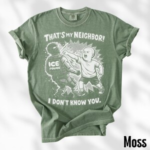 K&ouml;nnte beinhalten: Moosgr&uuml;nes T-Shirt mit einer grafischen Illustration eines Kindes, das einen ICE-Polizisten schl&auml;gt. Der Text auf dem Shirt lautet: "THAT'S MY NEIGHBOR! I DON'T KNOW YOU."