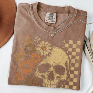May include: Brown Comfort Colors t-shirt featuring a floral and skull design. The graphic includes daisies and a checkered pattern. A silver necklace with a small pendant rests on the shirt. The shirt is size small.