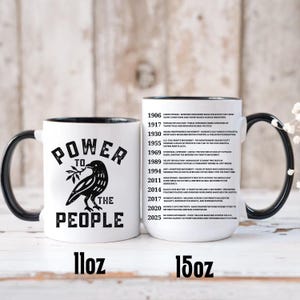 May include: White ceramic mug with a black handle and rim, featuring a black raven holding a sprig and the words "POWER TO THE PEOPLE." The mug is available in 11oz and 15oz sizes. A timeline of historical events is printed on the side.