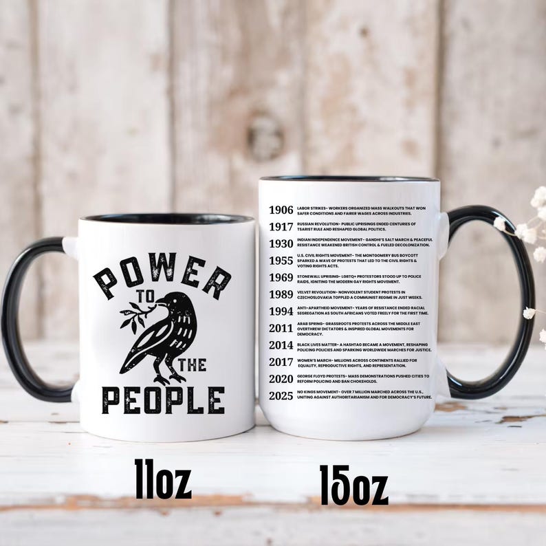 May include: White ceramic mug with a black handle and rim, featuring a black raven holding a sprig and the words "POWER TO THE PEOPLE." The mug is available in 11oz and 15oz sizes. A timeline of historical events is printed on the side.