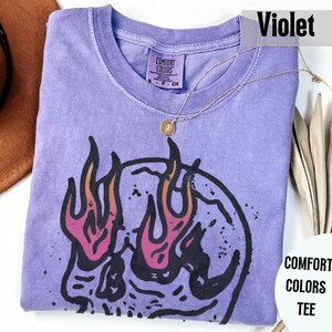 May include: Violet-colored Comfort Colors tee with a skull graphic and flames in pink and orange. The shirt has a crew neck and a small Comfort Colors tag. A gold necklace with a pendant is draped across the shirt.
