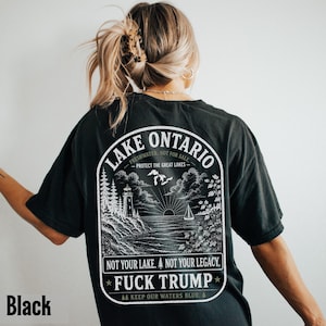 May include: A black t-shirt featuring a white graphic on the back. The design depicts a lighthouse, a sailboat, and a lake landscape with the text: LAKE ONTARIO, FRESHWATER, NOT FOR SALE, PROTECT THE GREAT LAKES, NOT YOUR LAKE. NOT YOUR LEGACY. FUCK TRUMP.