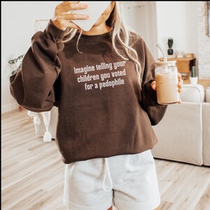 May include: A brown sweatshirt with the text "imagine telling your children you voted for a pedophile" in white. The person is also wearing gray shorts and holding a glass of iced coffee.