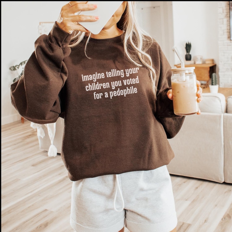 May include: A brown sweatshirt with the text "imagine telling your children you voted for a pedophile" in white. The person is also wearing gray shorts and holding a glass of iced coffee.