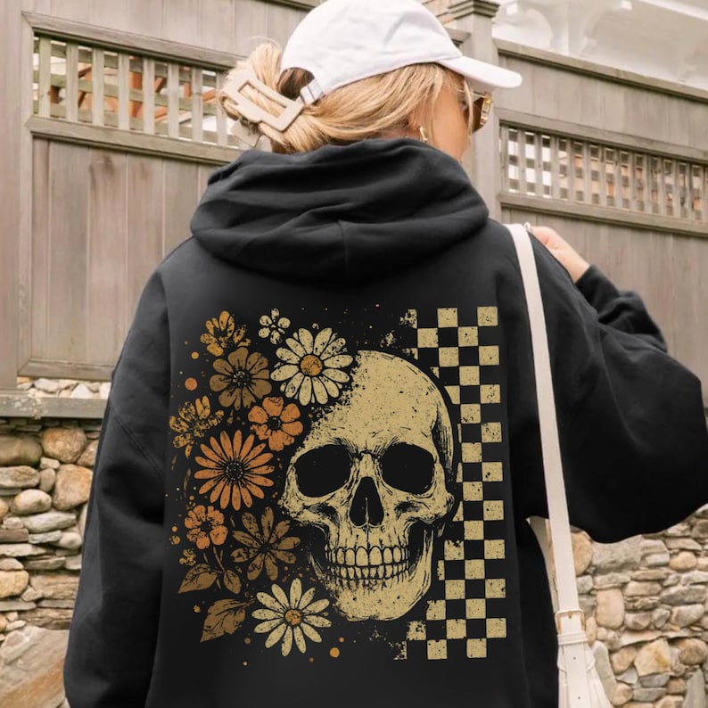 May include: Black hoodie featuring a skull design with floral accents in shades of orange and cream. A checkered pattern is on the right side of the skull. The hoodie is a casual garment.