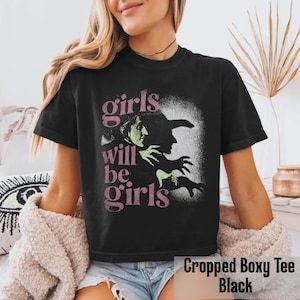 May include: A black short-sleeve t-shirt featuring a graphic print of a witch in profile with green skin and a black hat. The text "girls will be girls" is printed in a distressed pink font to the left of the witch illustration on the front.
