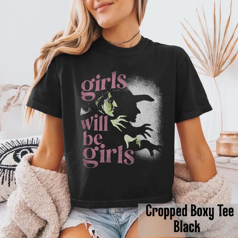 May include: A black short-sleeve t-shirt featuring a graphic print of a witch in profile with green skin and a black hat. The text "girls will be girls" is printed in a distressed pink font to the left of the witch illustration on the front.